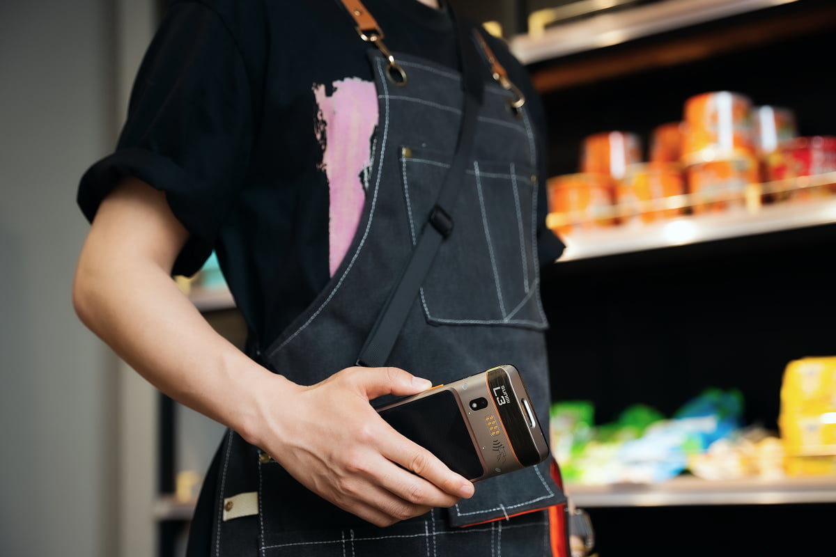 Retail staff member in an apron holding a Sunmi L3 handheld while working a stocked store shelf